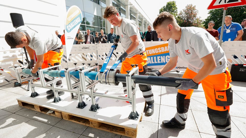 Drei Personen montieren im Freien ein Rohrsystem auf einer Metallkonstruktion bei einem Wettbewerb, im Hintergrund Zuschauer und ein Banner mit ‚Leitungsbau Challenge‘.