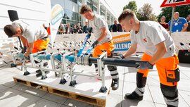 Drei Personen montieren im Freien ein Rohrsystem auf einer Metallkonstruktion bei einem Wettbewerb, im Hintergrund Zuschauer und ein Banner mit ‚Leitungsbau Challenge‘.