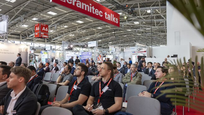 Ein Publikum sitzt auf Stühlen in der Messehalle A6 unter einem Schild auf dem 