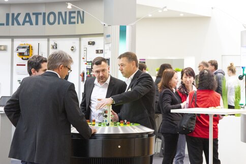 Four men in business attire stand around a round exhibition table on which various switches and buttons can be seen.