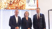 Three people in business attire stand smiling in front of an IFAT Munich key visual.