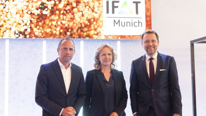 Three people in business attire stand smiling in front of an IFAT Munich key visual.