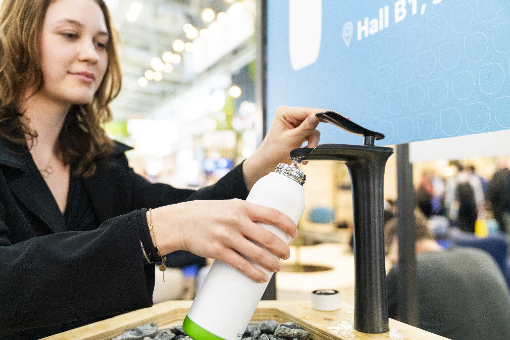 Junge Frau, die an einer Nachfüllstation in einer belebten Messehalle eine Wasserflasche auffüllt.
