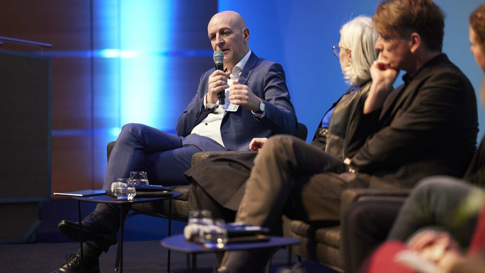 Ein Mann im Anzug, mit Glatze sitzt mit weiteren Personen auf einer Bühne und spricht in ein Mikrofon.