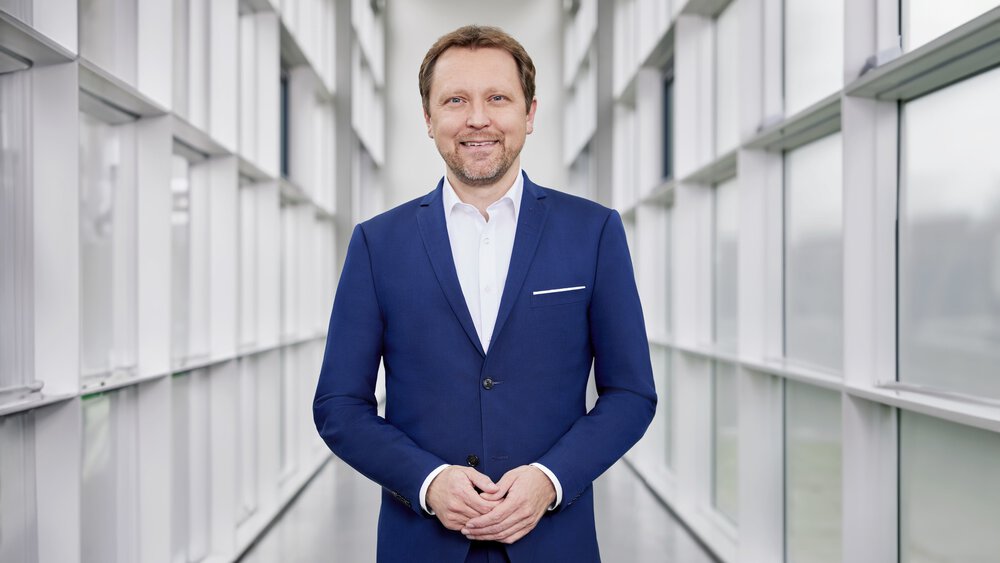 A man with short brown hair and a beard, wearing a blue suit and white shirt, stands smiling and symmetrically in a white hallway with many windows.