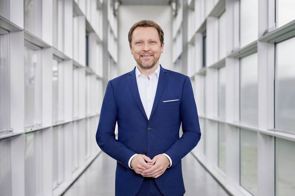 Ein Mann mit kurzen braunen Haaren und Bart in einem blauen Anzug mit weißem Oberhemd steht lächelnd und symmetrisch in einem weißen Gang mit vielen Fenstern.