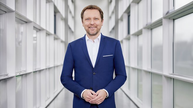 A man with short brown hair and a beard, wearing a blue suit and white shirt, stands smiling and symmetrically in a white hallway with many windows.