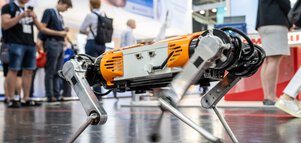 A four-legged orange and silver robot is exhibited at a technology fair, with people in the background.