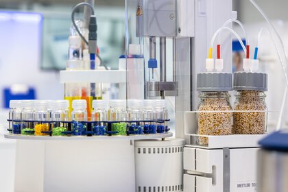 The picture shows a laboratory with various scientific instruments, colorful test tube racks and two large containers of grains.