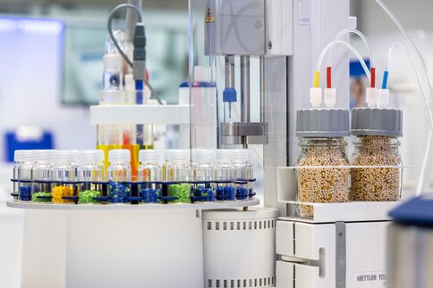 Das Bild zeigt ein Labor mit verschiedenen wissenschaftlichen Instrumenten, bunten Reagenzglasständern und zwei großen Behältern mit Körnern.