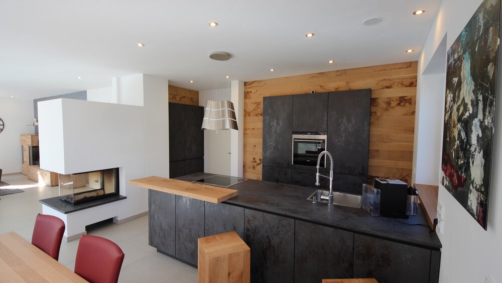 Blick in eine moderne schwarze Küche mit großer Arbeitsfläche, Backofen, Lampe, Kamin und einem Essbereich im Anschnitt.