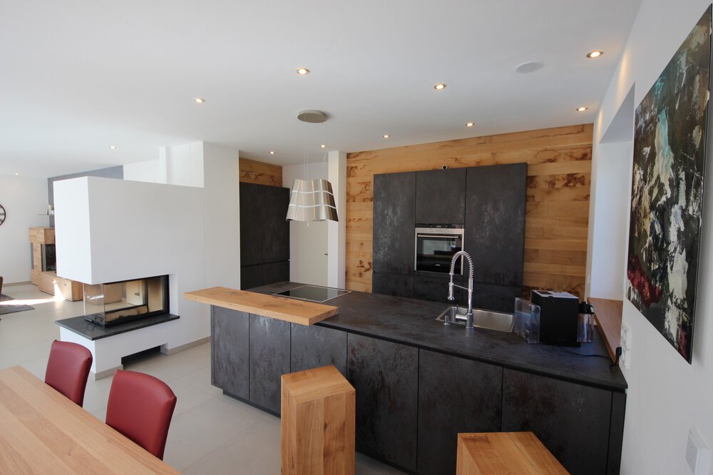 Blick in eine moderne schwarze Küche mit großer Arbeitsfläche, Backofen, Lampe, Kamin und einem Essbereich im Anschnitt.