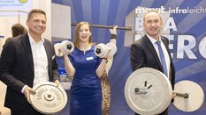 Auf der BAU stehen zwei Männer und eine Frau an einem Messestand und halten lächelnd verschiedene Hanteln und Gewichte in den Händen.