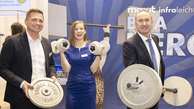 Two men and a woman are standing at a stand, smiling and holding various dumbbells and weights in their hands, which appear to be made of lightweight concrete.