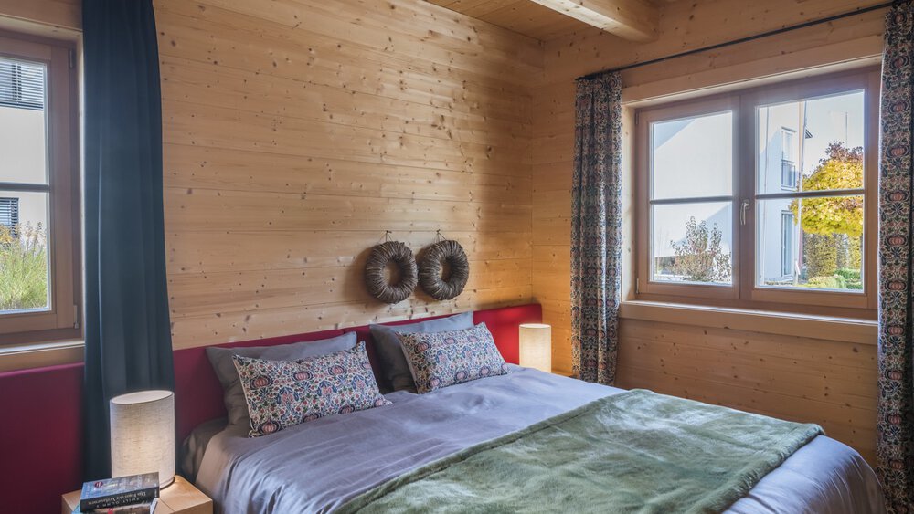 Gemütliches Schlafzimmer mit einem Bett, bunten Kissen, einer Lampe und Sonnenlicht, das durch ein Fenster mit Vorhängen strömt.