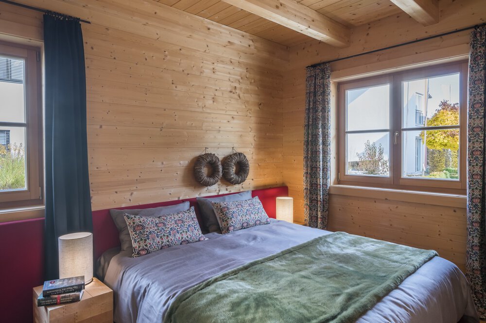 Gemütliches Schlafzimmer mit einem Bett, bunten Kissen, einer Lampe und Sonnenlicht, das durch ein Fenster mit Vorhängen strömt.