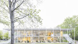 Modernes zweistöckiges Glasgebäude, umgeben von Bäumen, mit grasbewachsenem Vordergrund und einer Holzbank.