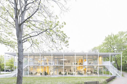 Modernes zweistöckiges Glasgebäude, umgeben von Bäumen, mit grasbewachsenem Vordergrund und einer Holzbank.