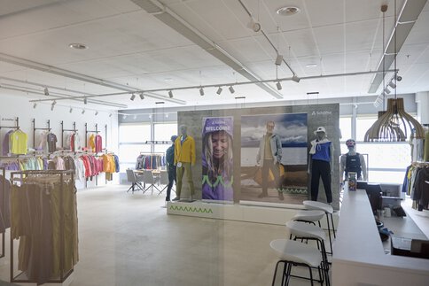 Inneneinrichtung des Schöffel Showrooms im MOC Ordercenter mit farbenfroher Kleidung auf Ständern und Schaufensterpuppen neben einer Theke mit Hockern.