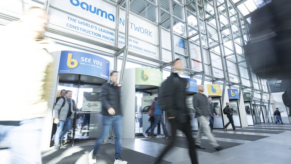 Besucher betreten das Messegelände durch Drehtüren, Glasfassade und Hinweisschilder auf die bauma 2028 sichtbar.