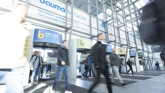 Besucher betreten das Messegelände durch Drehtüren, Glasfassade und Hinweisschilder auf die bauma 2028 sichtbar.