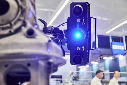 Close-up of a 3D scanner with blue light scanning a mechanical component. Trade fair stands can be seen in the background.