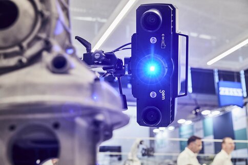 Close-up of a 3D scanner with blue light scanning a mechanical component. Trade fair stands can be seen in the background.