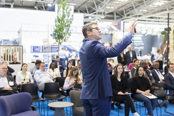 Mann im blauen Anzug spricht vor einem sitzenden Publikum in einer Veranstaltungshalle.
