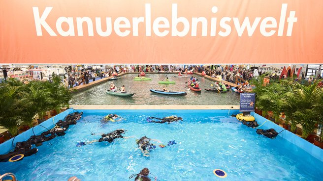 Indoor-Wasserbecken auf der Messe f.re.e mit Tauchern und Kanufahrern unter dem Banner „Kanuerlebniswelt“, umgeben von Besuchern und Pflanzen.