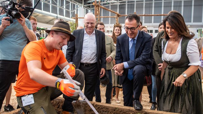 A man kneels in a raised bed in an exhibition hall, surrounded by spectators, while a cameraman films over his shoulder.