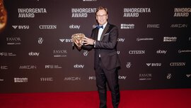 A smiling man with short hair, glasses and wearing a suit stands in front of a photo wall of the INHORGENTA AWARD and holds a trophy.