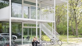Person, die an einem modernen zweistöckigen Gebäude vorbeigeht, vor dem Fahrräder geparkt sind und im Hintergrund Bäume zu sehen sind.