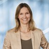 Portrait of a person with shoulder-length hair wearing a beige blazer in front of a blurred blue background with graphic lines.