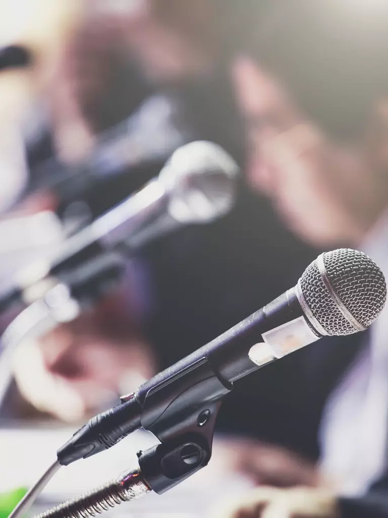 Close-up of microphones at a press conference with blurred figures in the background.