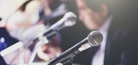 Nahaufnahme von Mikrofonen bei einer Pressekonferenz mit unscharfen Figuren im Hintergrund.