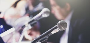 Close-up of microphones at a press conference with blurred figures in the background.