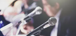 Ein Mikrofon steht im Vordergrund, während mehrere Mikrofone und Personen im Hintergrund sichtbar sind. Das Bild suggeriert eine Pressekonferenz.