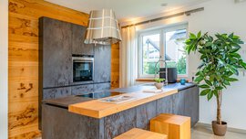 Moderne Küche mit Oberflächen aus Holz und Metall, einer Kücheninsel mit Hockern, einer Topfpflanze, einem Backofen, einem großen Fenster und einer Pendelleuchte.