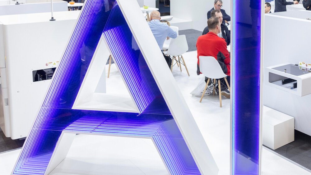 Large glowing AI letters in blue stand on a trade fair floor while visitors in the background sit at white tables having conversations