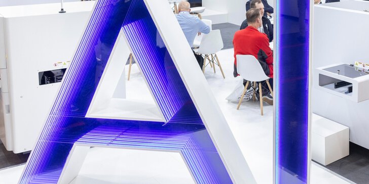 Large glowing AI letters in blue stand on a trade fair floor while visitors in the background sit at white tables having conversations