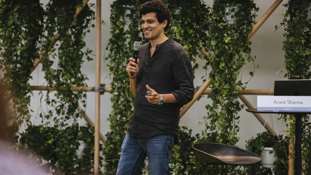 Person auf einer Bühne, die in ein Mikrofon spricht, daneben ein Stuhl und ein Schild, auf dem „Anant Sharma“ steht.