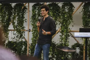 Person on a stage speaking into a microphone, next to a chair and a sign saying ‘Anant Sharma’.