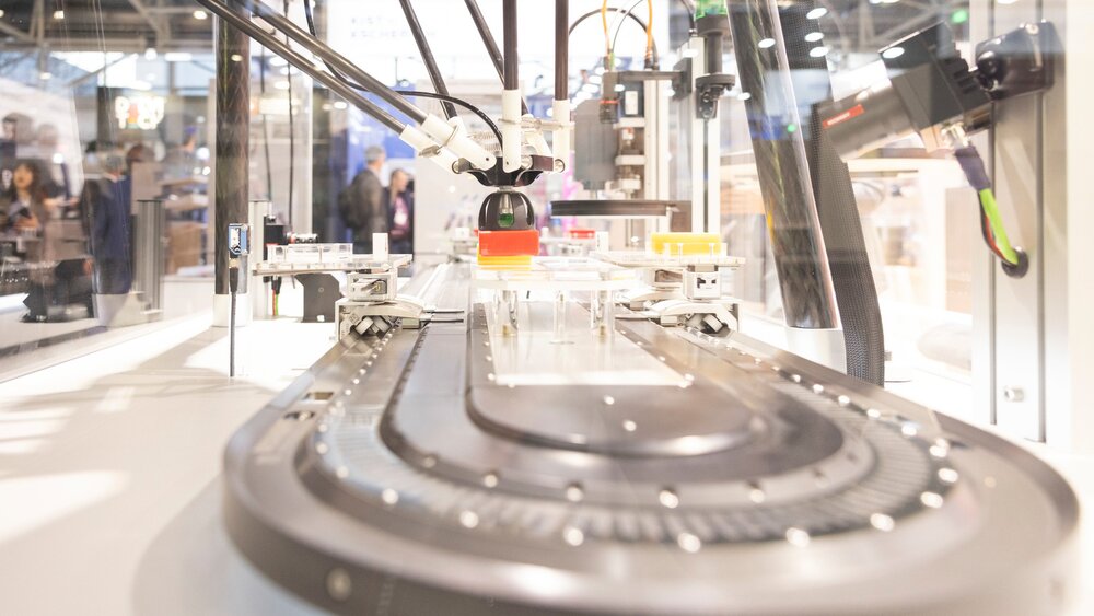 Eine durchsichtige Maschine mit Roboterarmen und einer Transportvorrichtung steht an einem Stand auf der productronica.