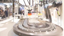 Eine durchsichtige Maschine mit Roboterarmen und einer Transportvorrichtung steht an einem Stand auf der productronica.