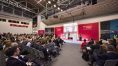 Zahlreiche Messebesucher sitzen auf Stühlen vor einer Bühne in einer Halle der transport logistic und sehen sich einen Vortrag von zwei Männern an.