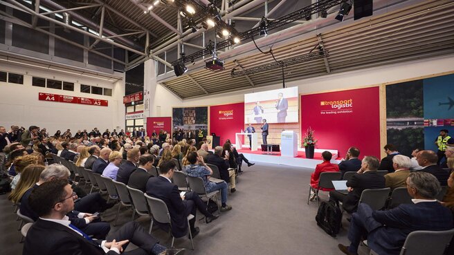 Zahlreiche Messebesucher sitzen auf Stühlen vor einer Bühne in einer Halle der transport logistic und sehen sich einen Vortrag von zwei Männern an.
