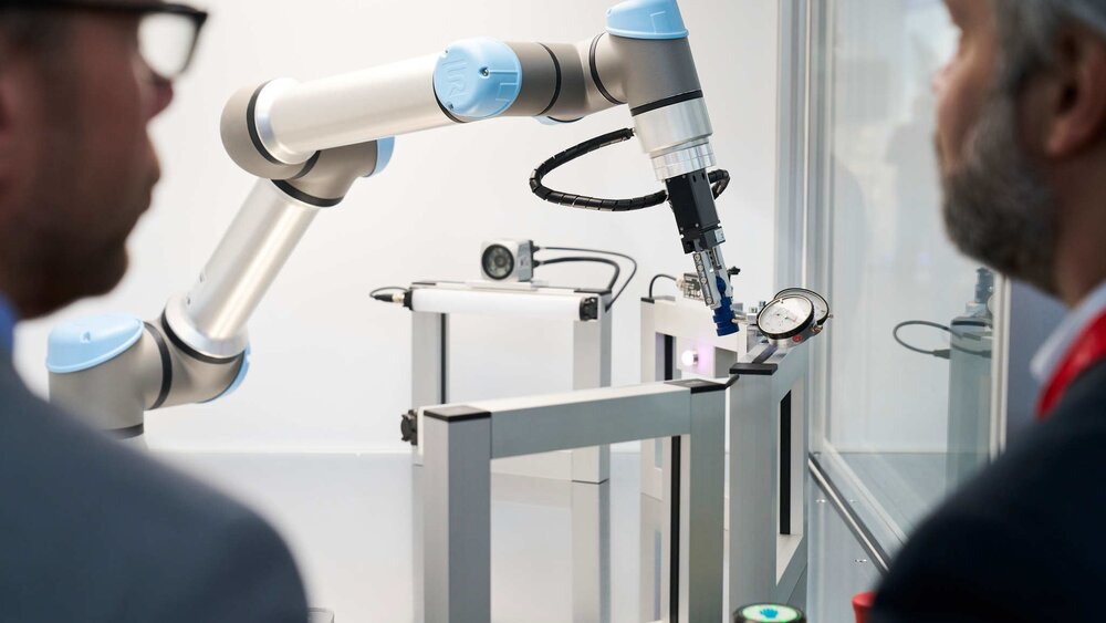 Blick über die Schultern zweier Männer auf einen weiß-grau-blauen Roboterarm mit mehreren viereckigen weißen Rahmen daneben.