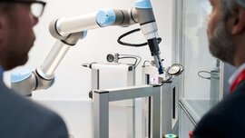 Blick über die Schultern zweier Männer auf einen weiß-grau-blauen Roboterarm mit mehreren viereckigen weißen Rahmen daneben.