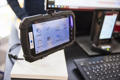 A robust-looking, industrial navigation device on a support arm, which is switched on and displayed at a trade fair stand. There is a computer in the background.