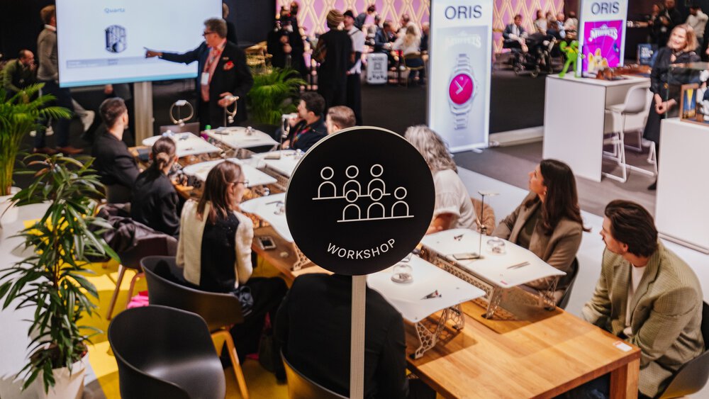 A round black sign with six stylised figures and the word “WORKSHOP” underneath. Behind it, several people are sitting at jewellery tables.